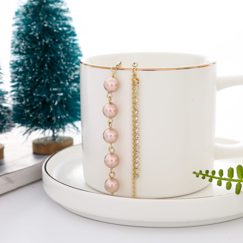 Aretes asimétricos dorados con perlas rosadas colgando del borde de una taza