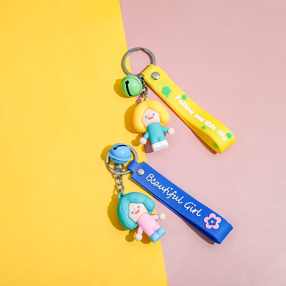 Dos llaveros con figuras de muñequitas de cabello corto, correas en azul y amarillo con frases y cascabeles de colores